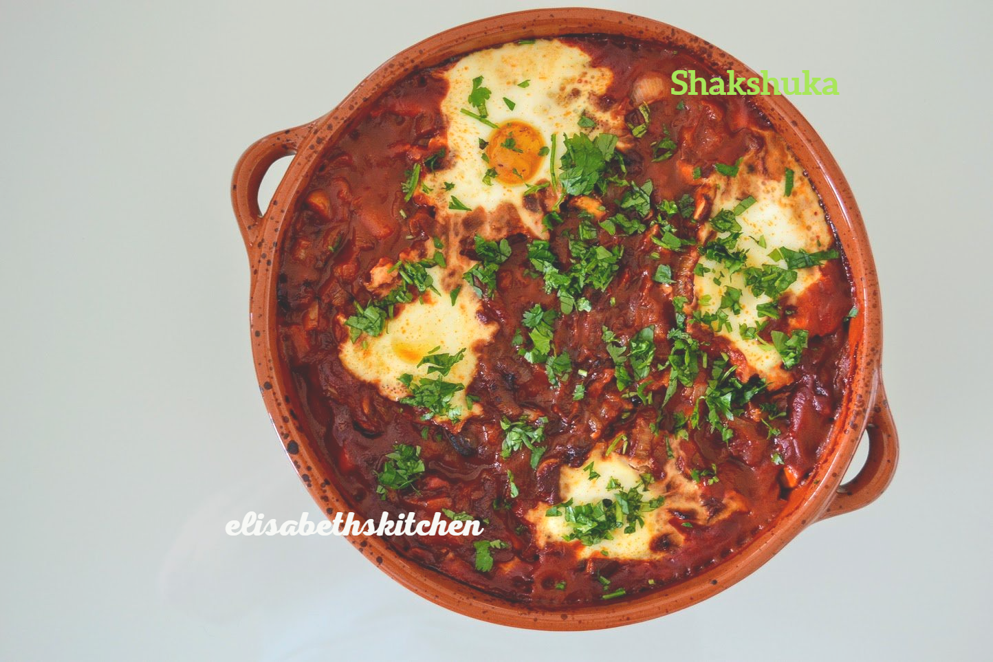 Shakshuka, een stoofpot van tomaat en paprika met gepocheerde eieren, geserveerd in een aardewerken schaal en gegarneerd met peterselie.