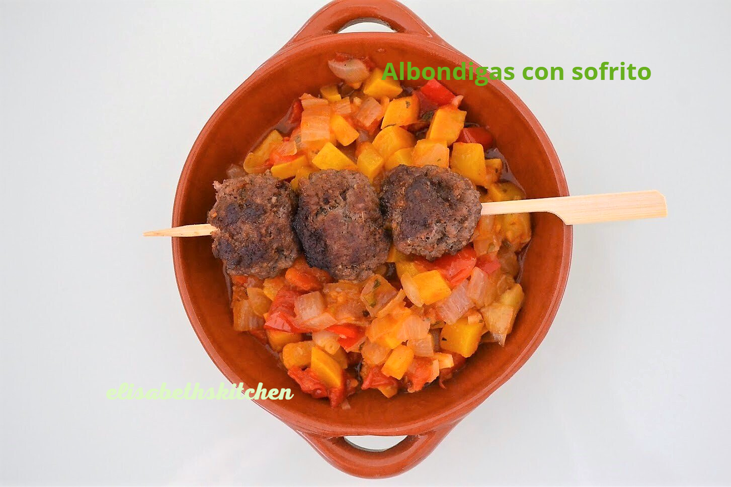 Gehaktballetjes aan een spiesje boven een sofrito van groenten in een terracotta kom.