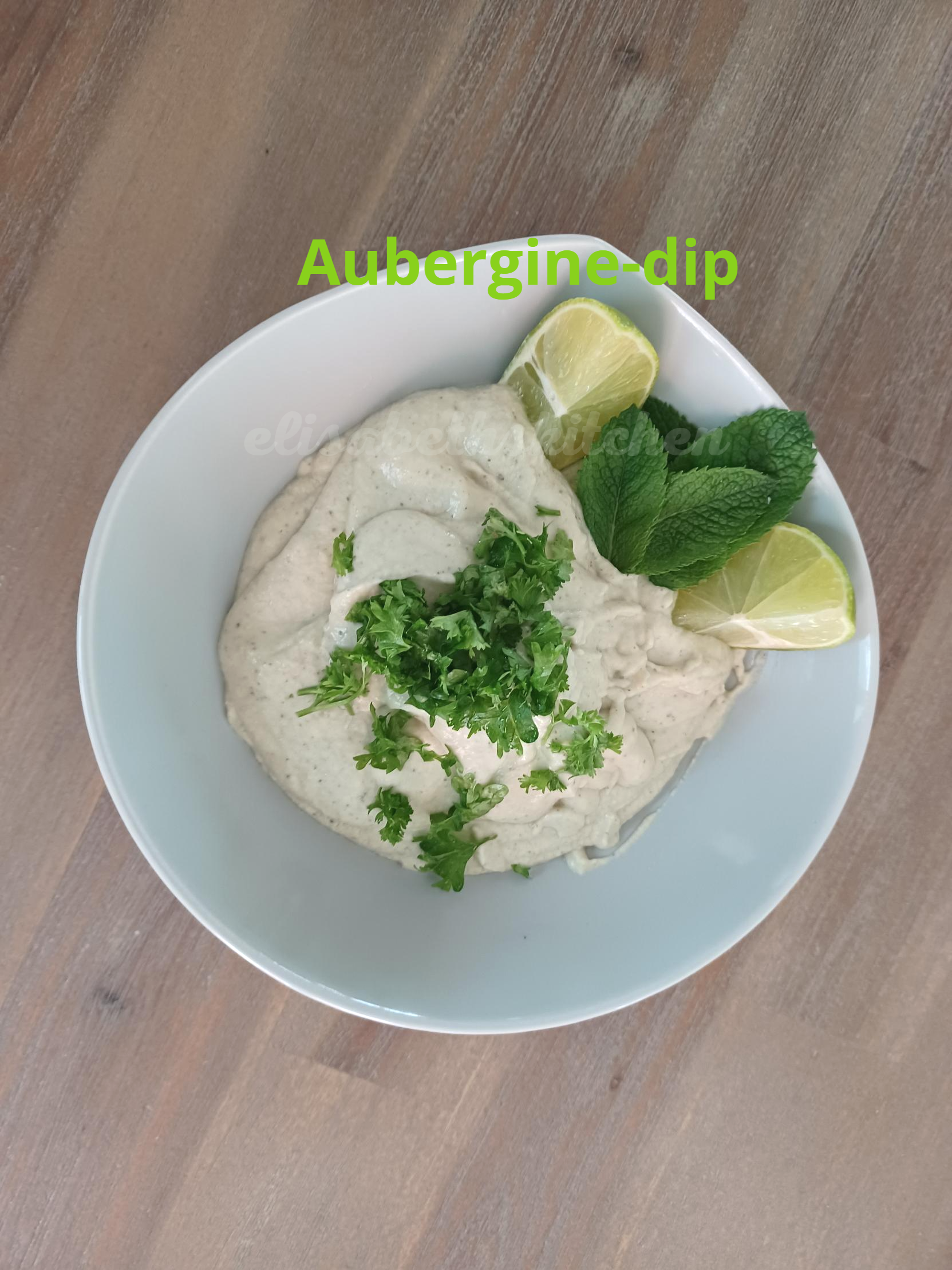 Verderop de pagina staat het recept Auberginesaus in een kom, gegarneerd met peterselie, munt en partjes limoen, op een houten ondergrond.