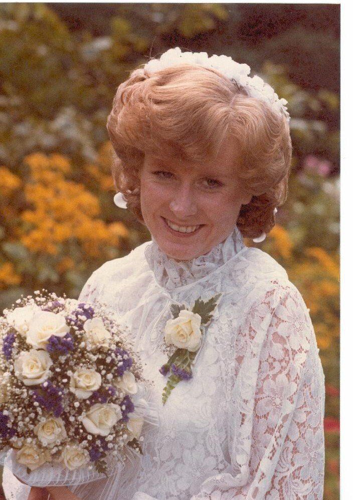 Vrouw in een witte kanten trouwjurk met bloemen in haar hand, lachend in een tuin.