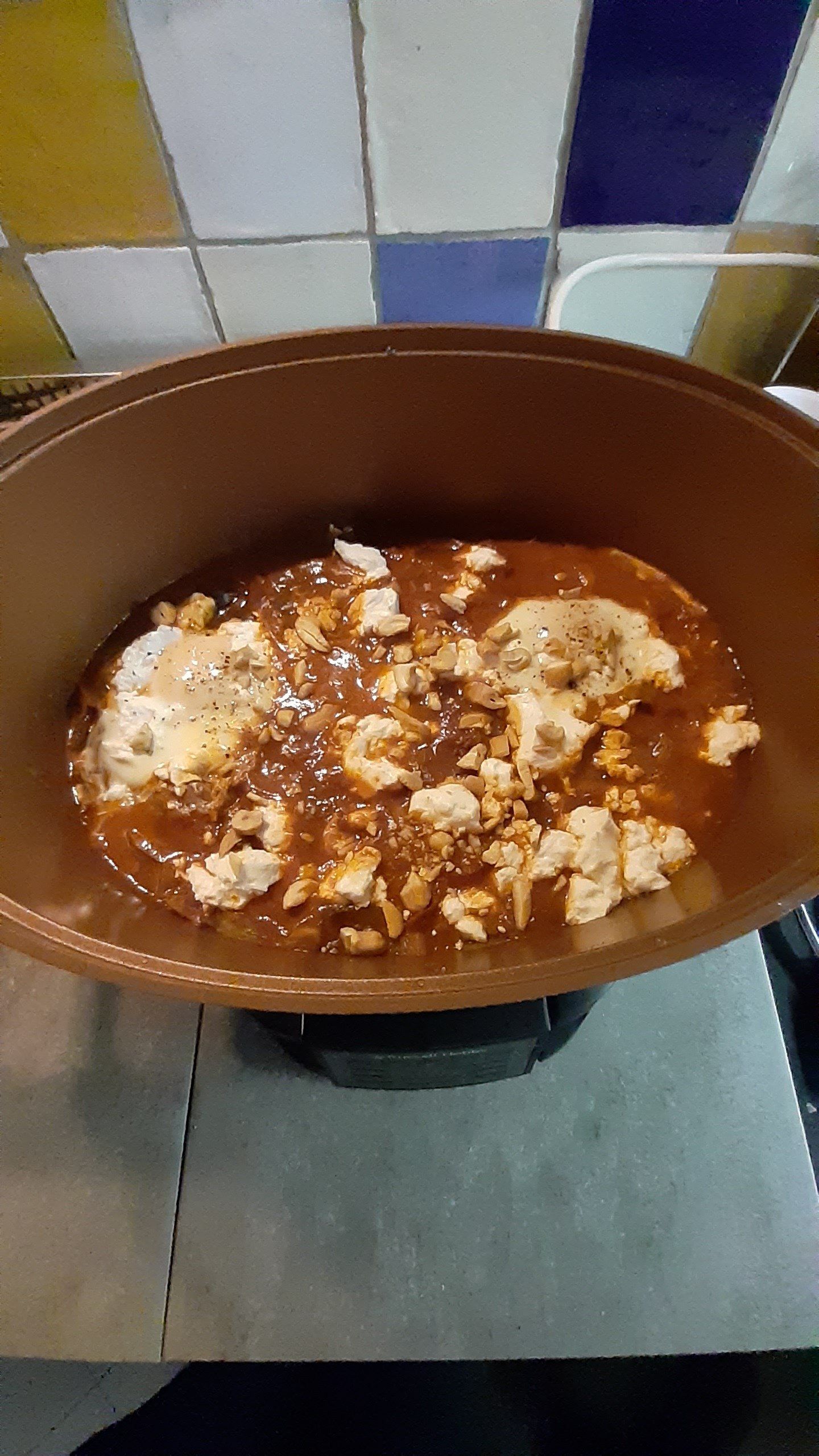 Een bruine ovenschaal gevuld met een sausrijk gerecht, bedekt met witte kruimels, tegen een betegelde achterwand.