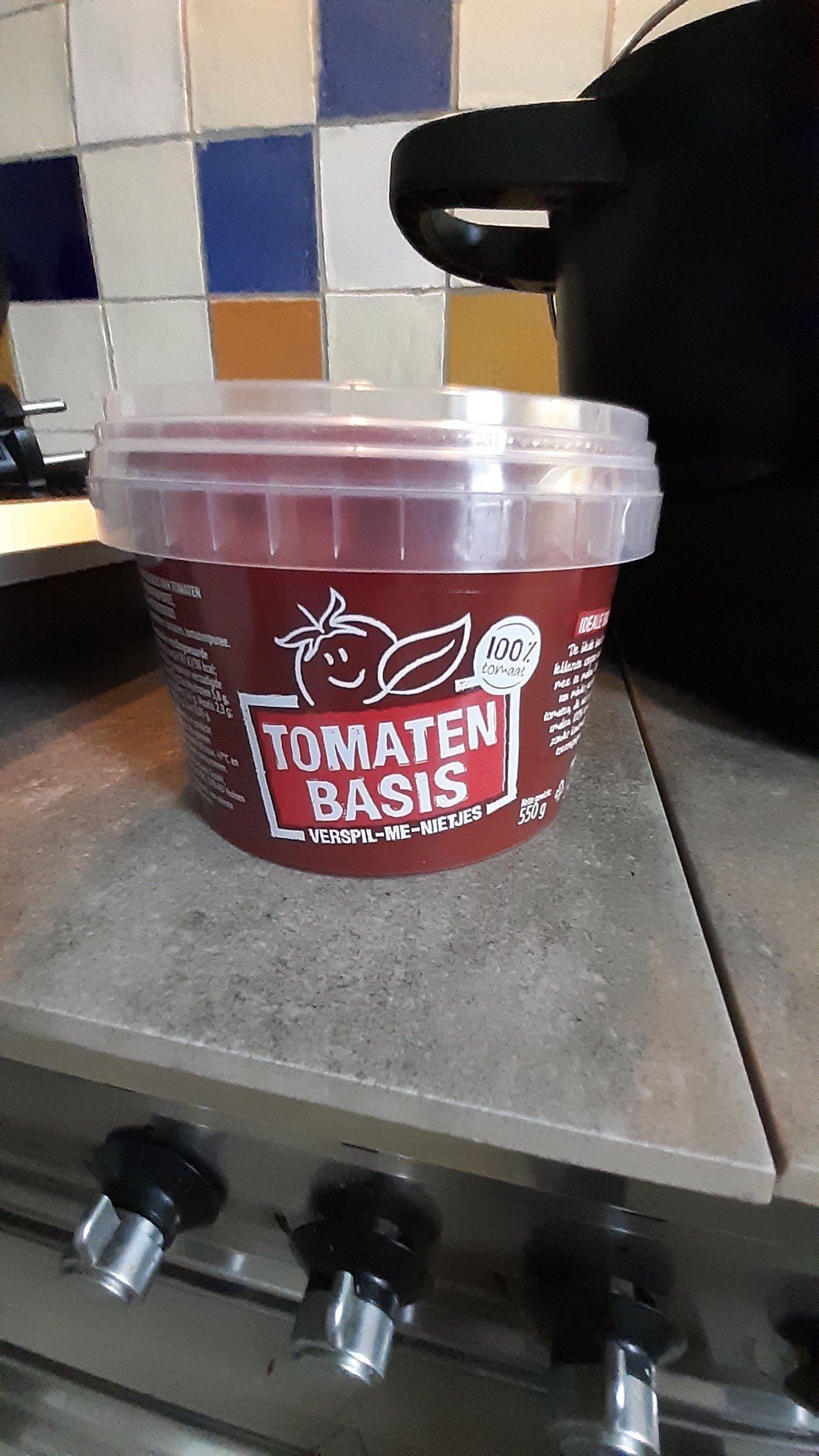 Een bakje "Tomaten Basis" tomatensaus staat op het fornuis, naast een zwarte pan en een betegelde achterwand.
