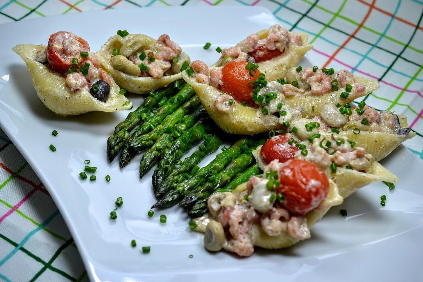 Pastaschelpen gevuld met garnalen, tomaten en kruiden, geserveerd met asperges op een wit bord.