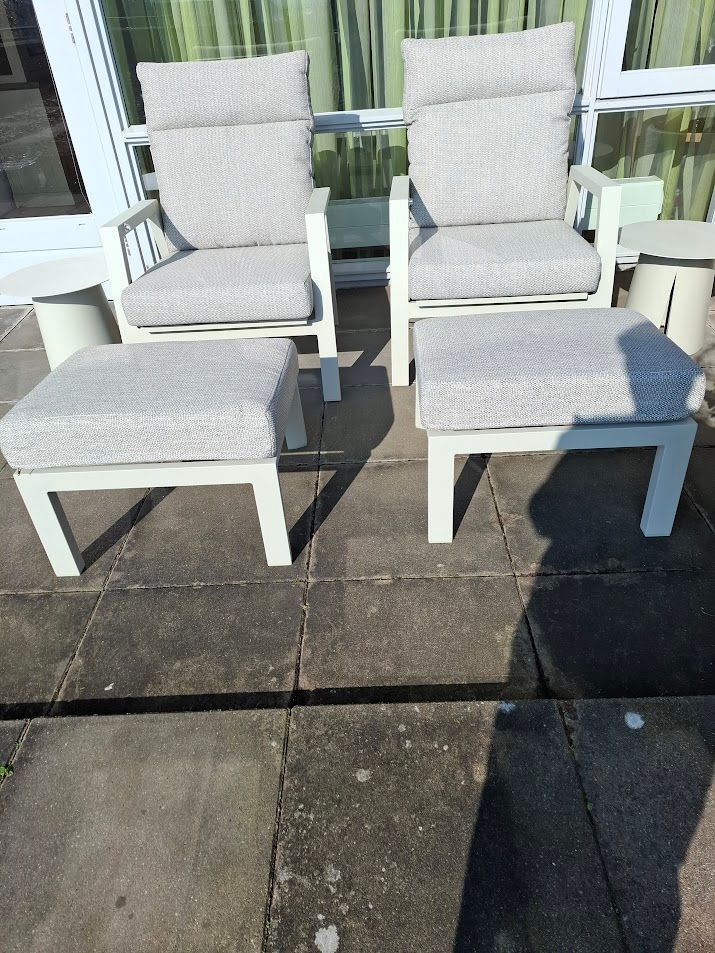 Twee witte buitenstoelen met grijze kussens en bijpassende voetenbankjes op een terras.