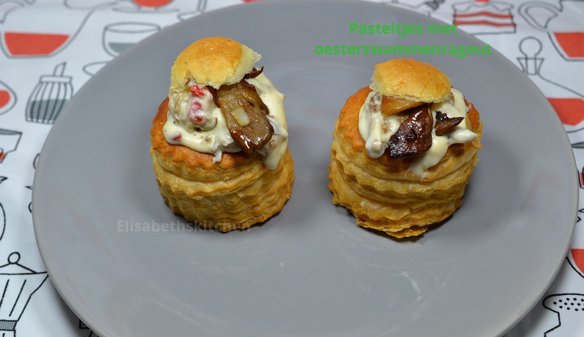 Twee bladerdeeggebakjes gevuld met romige saus, champignons en een klein champignonhoedje op een grijs bord.