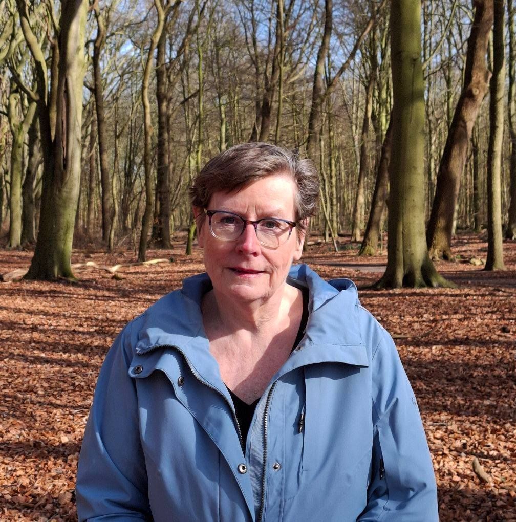 Een vrouw met een bril en een blauwe jas staat in een bos met kale bomen en gevallen bladeren.