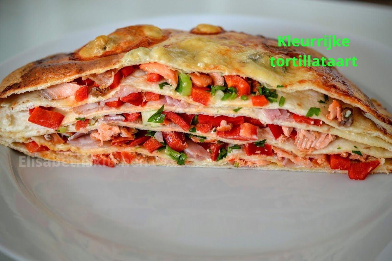 Een kleurrijke, gelaagde tortillacake op een wit bord, gevuld met zalm, rode paprika en groene bladgroenten.