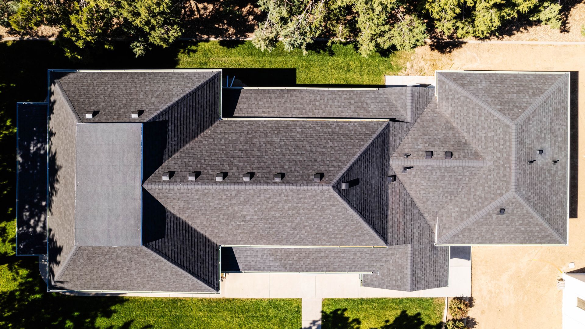 Overhead view of a dark grey shingle roof on a house, with trees and green grass surrounding it.