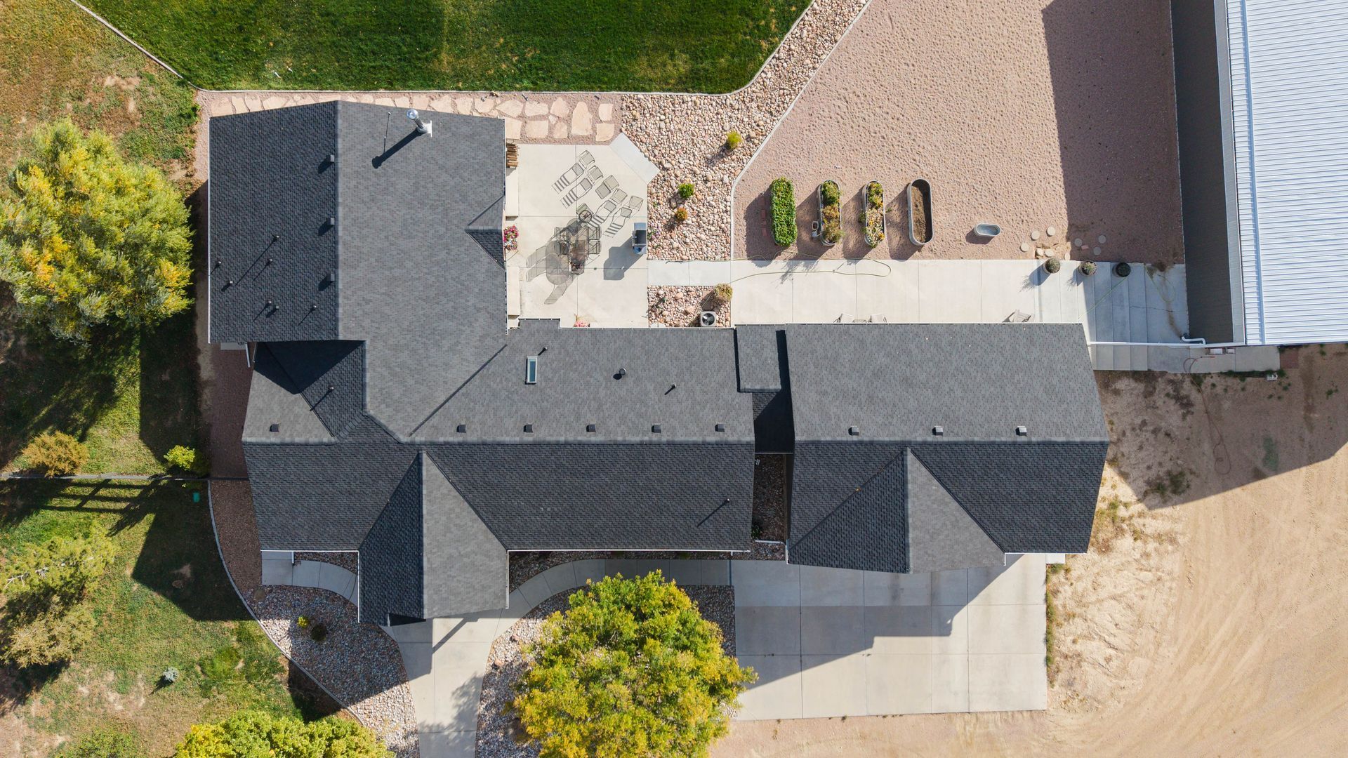 Overhead view of a dark-roofed house with a concrete driveway, patio, and surrounding dry landscape and grass.