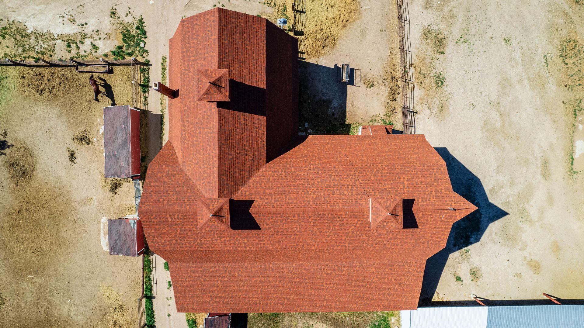 Overhead view of a multi-roofed reddish-brown building complex on dirt, with a hint of green grass.