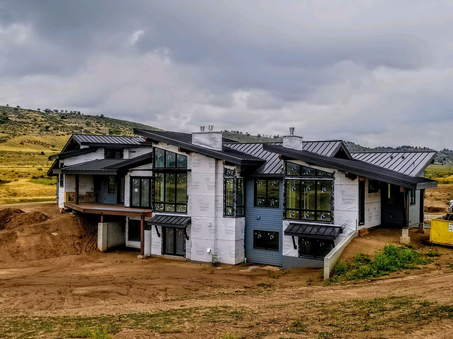 Modern house under construction with gray siding, black roof, and large windows on a hillside.