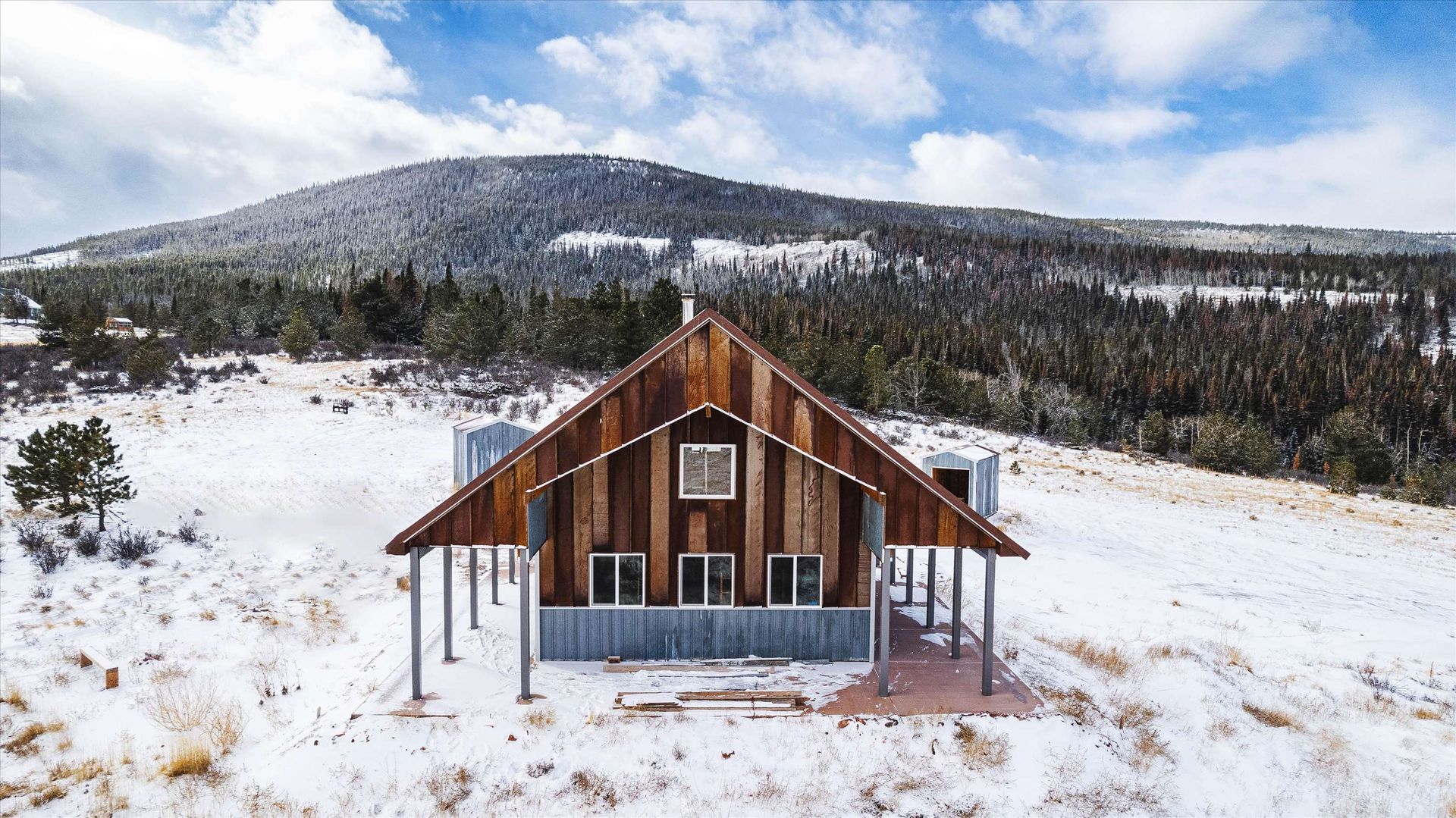 Wooden cabin in snowy field with a mountain in the background, under a blue sky.