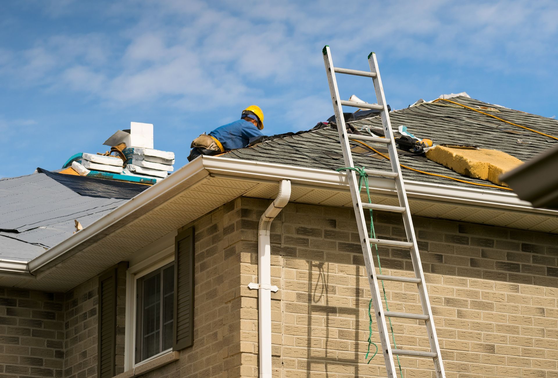 two construction workers repairing a roof on a brick house under a partly cloudy sky.