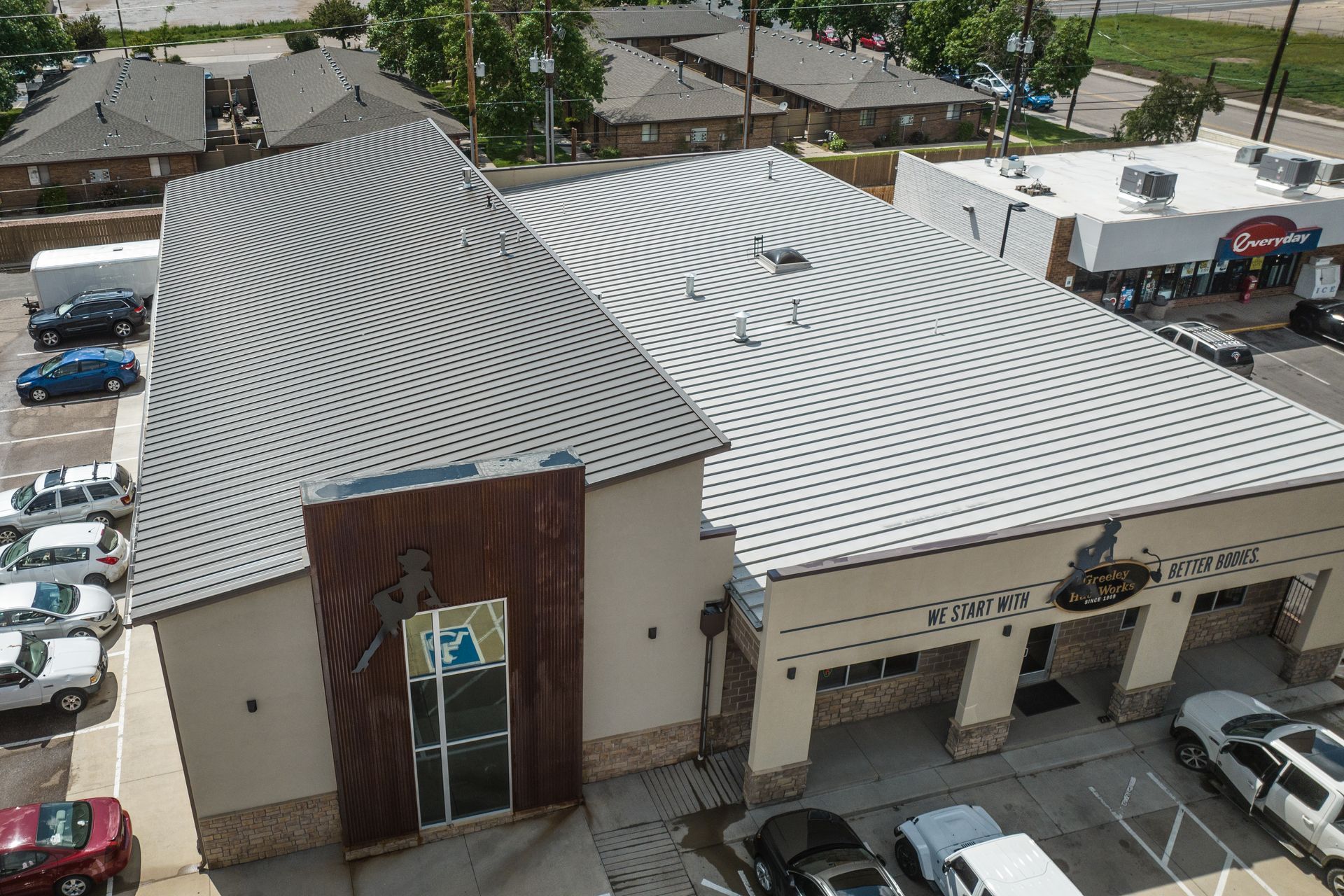 Aerial view of a building with a uniquely curved metal roof, parking lot, and other buildings.