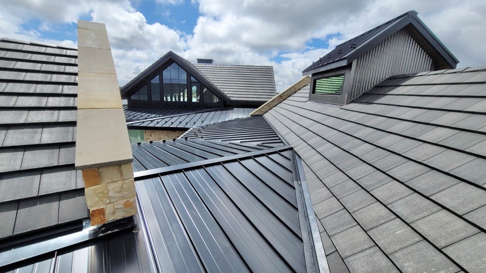 Multi-level roof with dark shingles and metal panels, chimney, and partially cloudy sky.