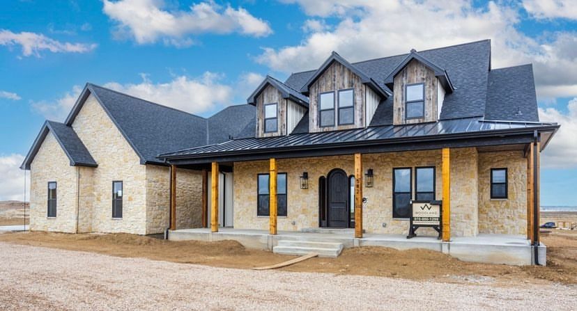 Beige brick farmhouse with black roof, wooden porch, and three dormer windows.