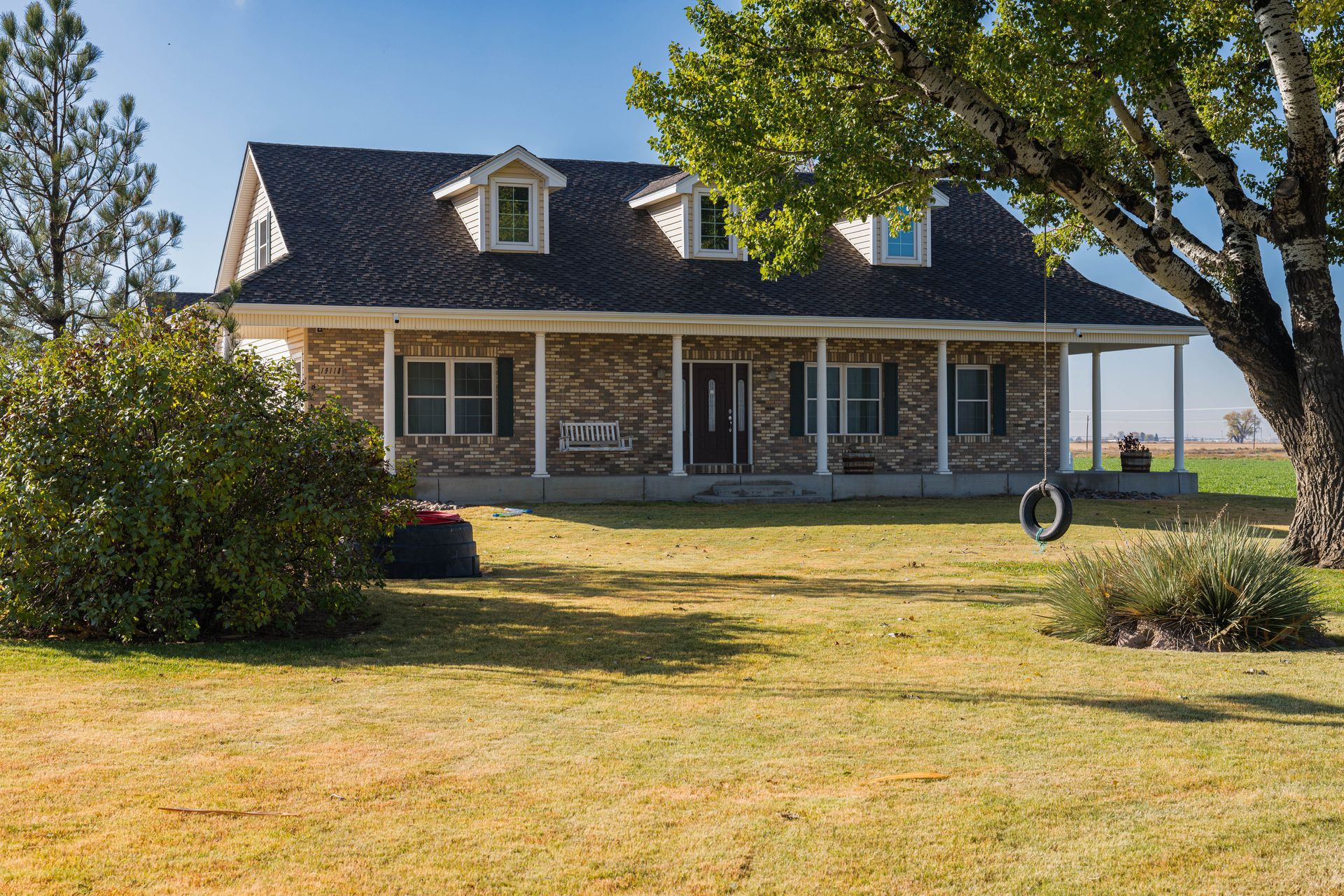 House with a porch, brick exterior, and dormer windows on a large grassy lawn, with a tire swing hanging from a tree.