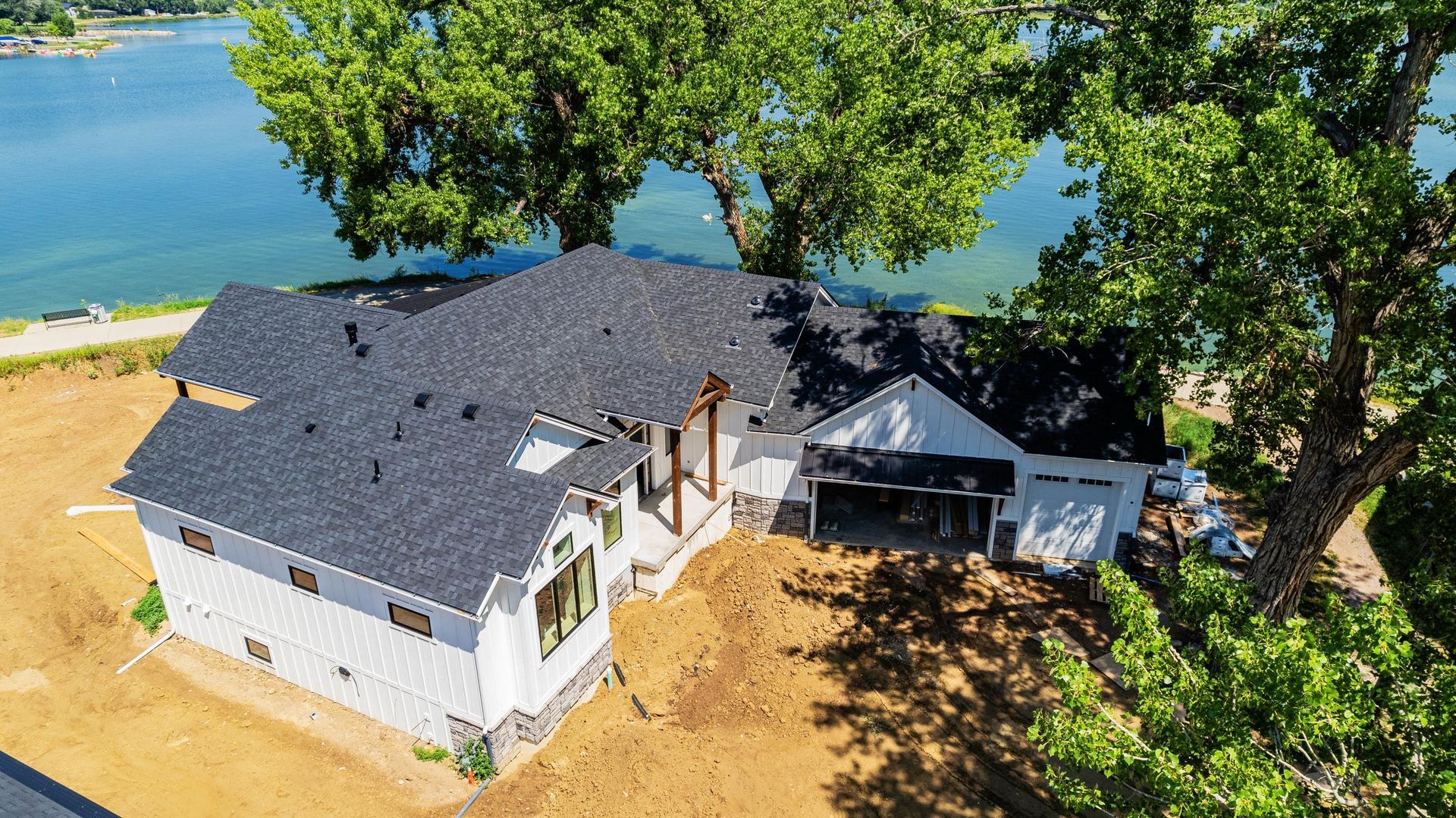White house with black roof, near a lake, with a large tree.