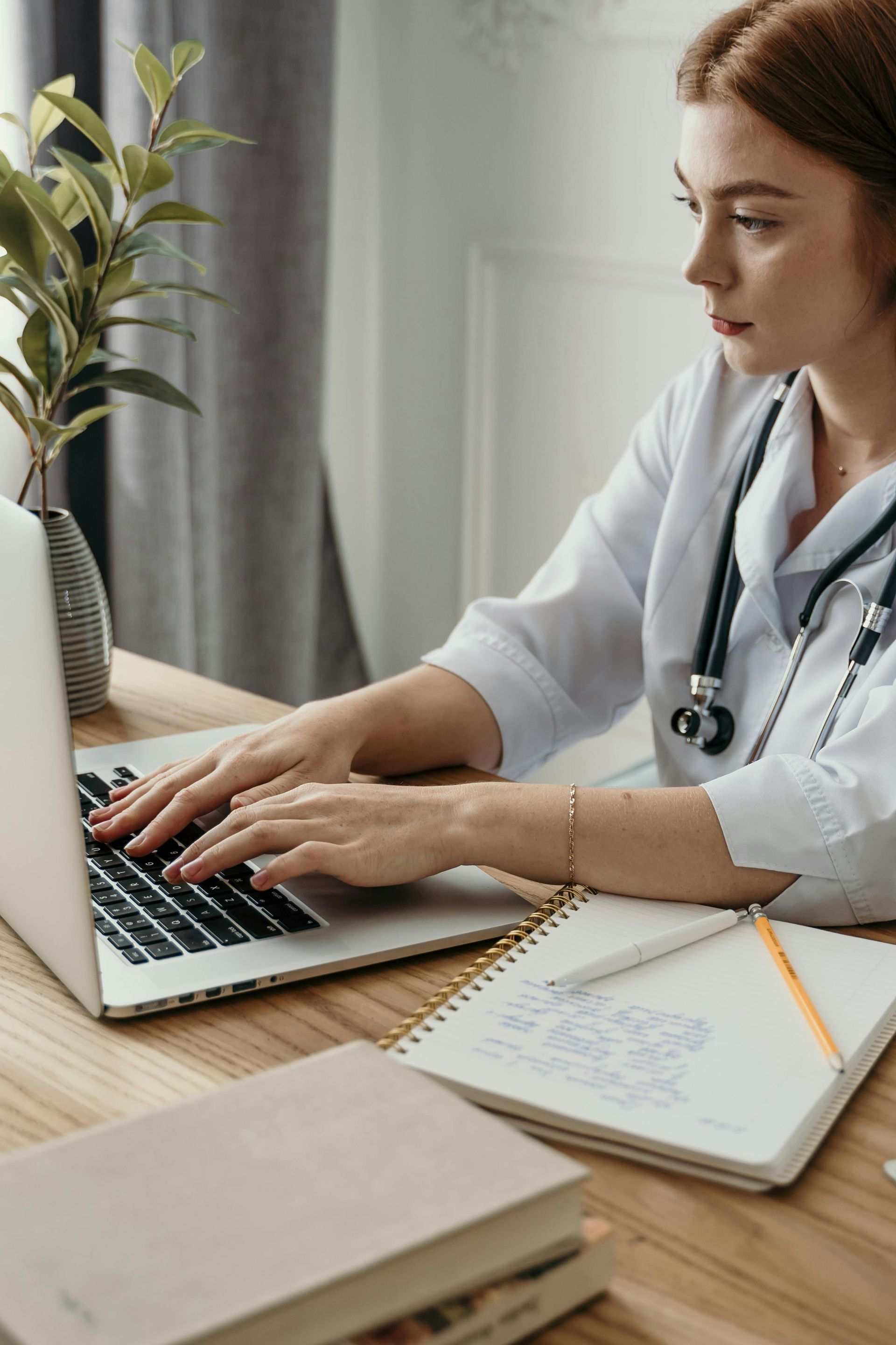 Woman in white coat, stethoscope around neck, typing on laptop at wooden desk with notebook and plant.