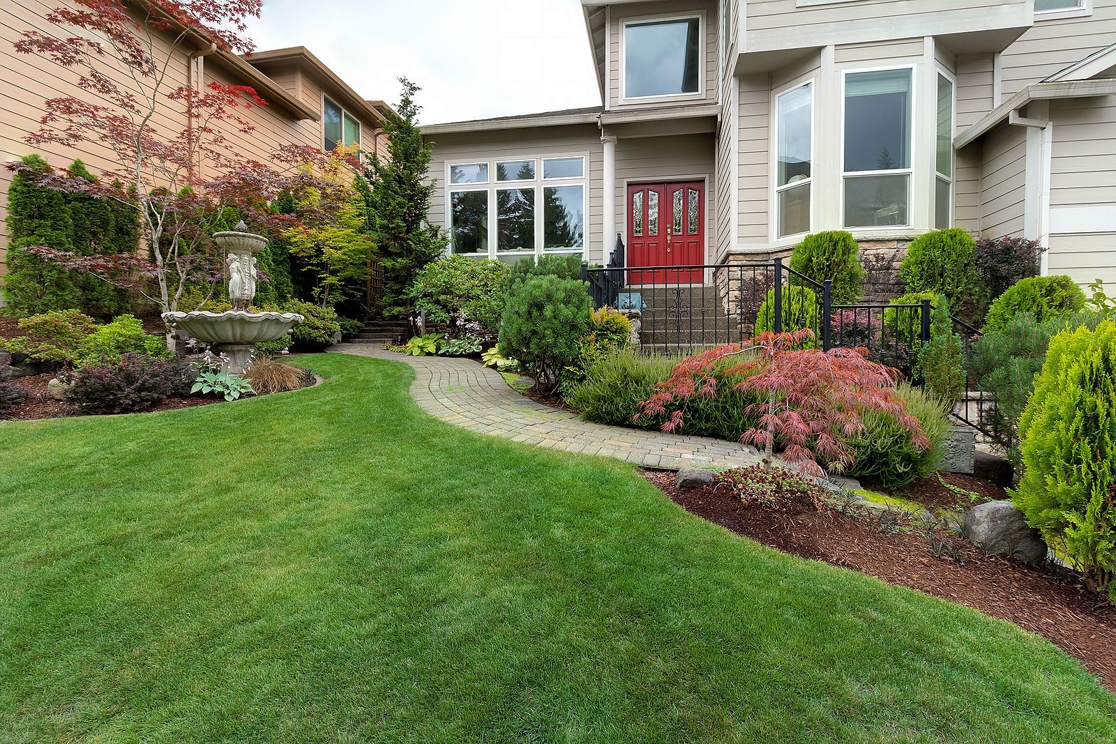 A manicured front lawn leads to a stone walkway and a house with a red front door, surrounded by shrubs and trees.