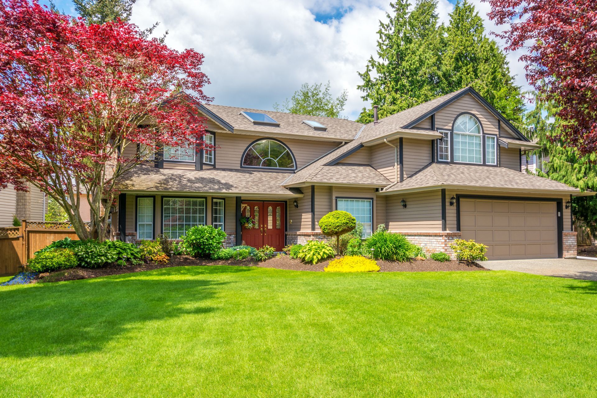 A two-story suburban home with tan siding, a red front door, and a manicured green lawn, framed by lush trees.