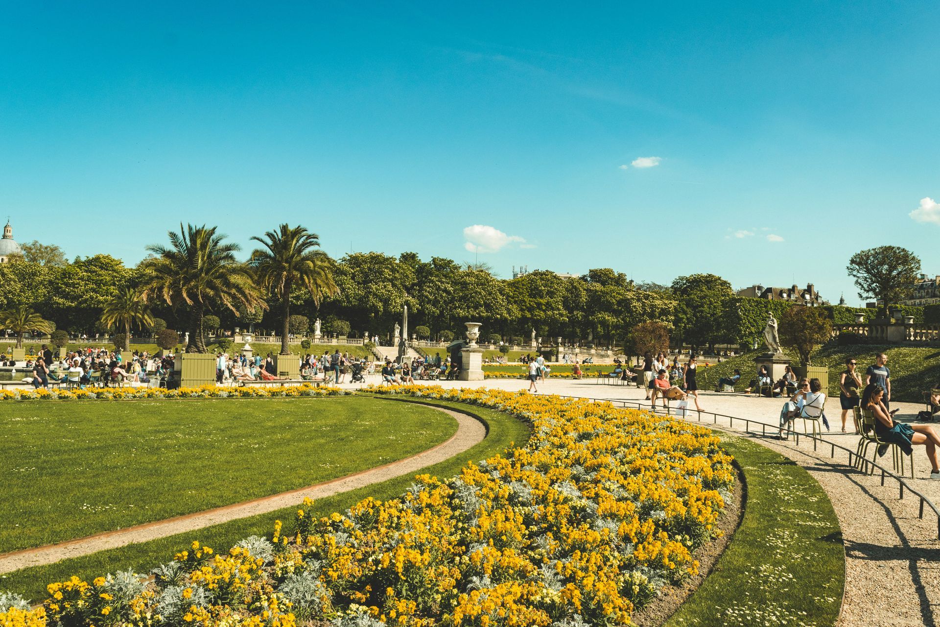 A sunny day in a Parisian garden with a bright yellow flower bed in the foreground, green grass, and people sitting.