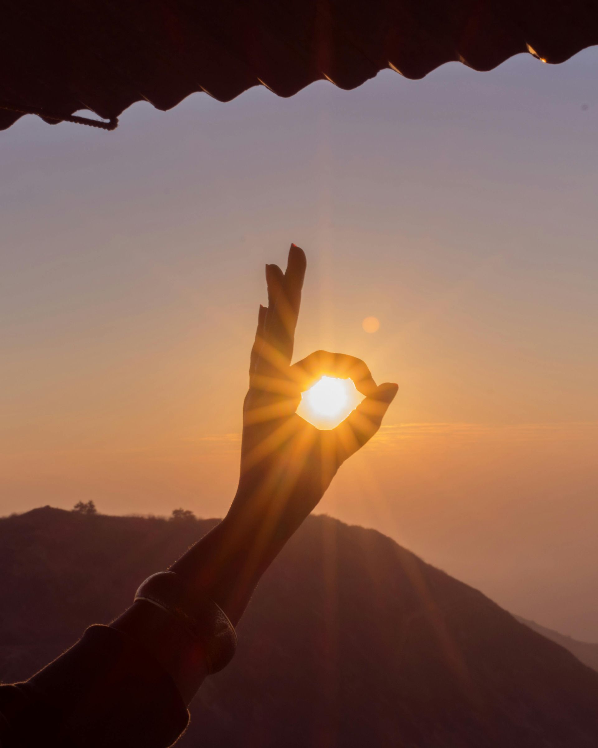 Hand making an OK sign to frame the sun at sunset over mountains