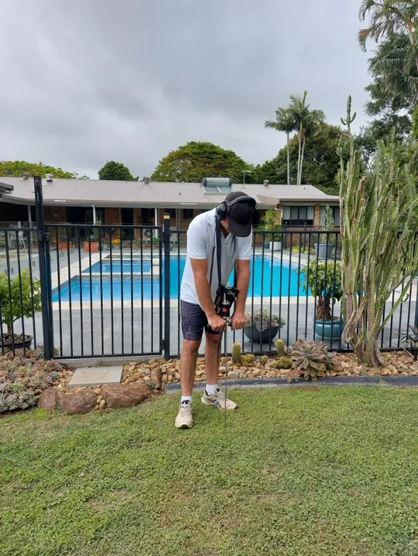Man using detection equipment in a grassy yard, with a pool in the background. Cloudy sky.