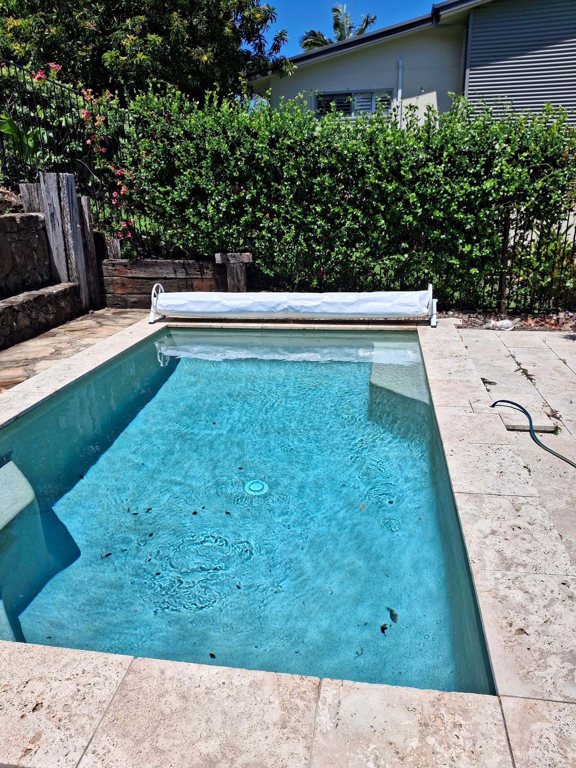 A rectangular swimming pool with blue water and a white cover, surrounded by stone and greenery.