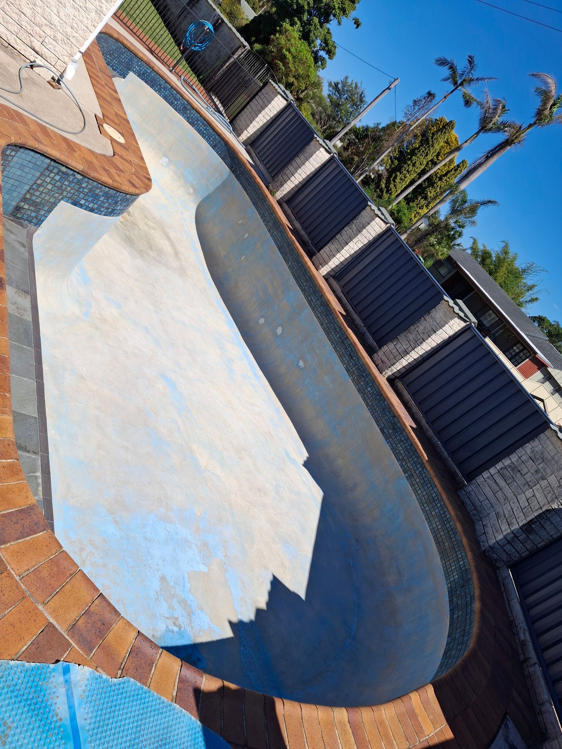 Empty, kidney-shaped pool with blue and brown tiling, surrounded by brick and a fence, under a blue sky.