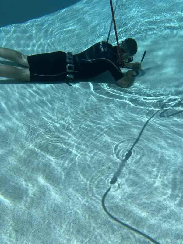 Scuba diver using a tool underwater, wearing a wetsuit, over a sandy bottom in a pool.