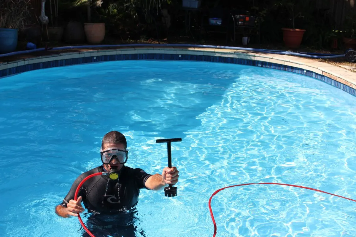 Diver in pool holding a cleaning tool; sunny day, blue water.