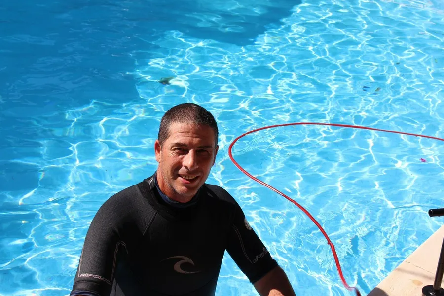 Man in a wetsuit by a pool. He’s smiling. Bright blue water. Red rope.