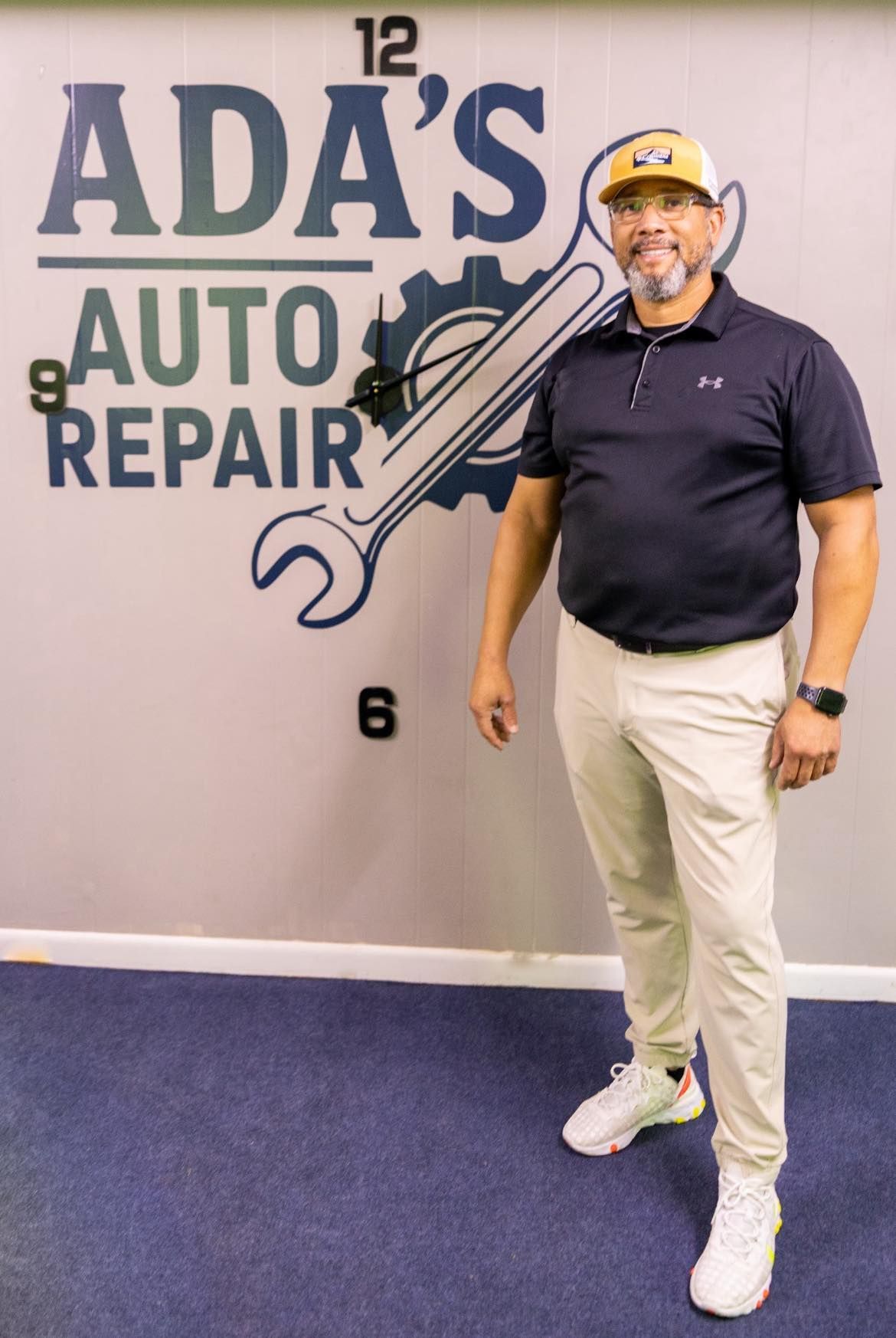 Man standing in front of Ada's Auto Repair logo on a wall. He's wearing a polo shirt, pants, and a baseball cap.