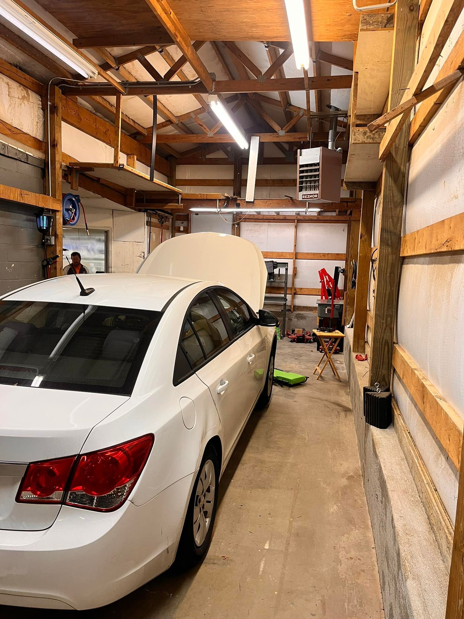 White car with hood up in a garage, under exposed wood beams and lighting.