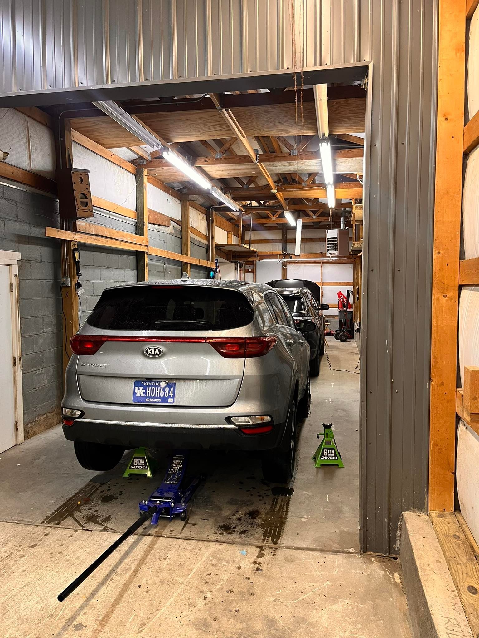 Silver Kia SUV in a garage bay, lifted with jack and stands. Other cars, tools, and lights visible.