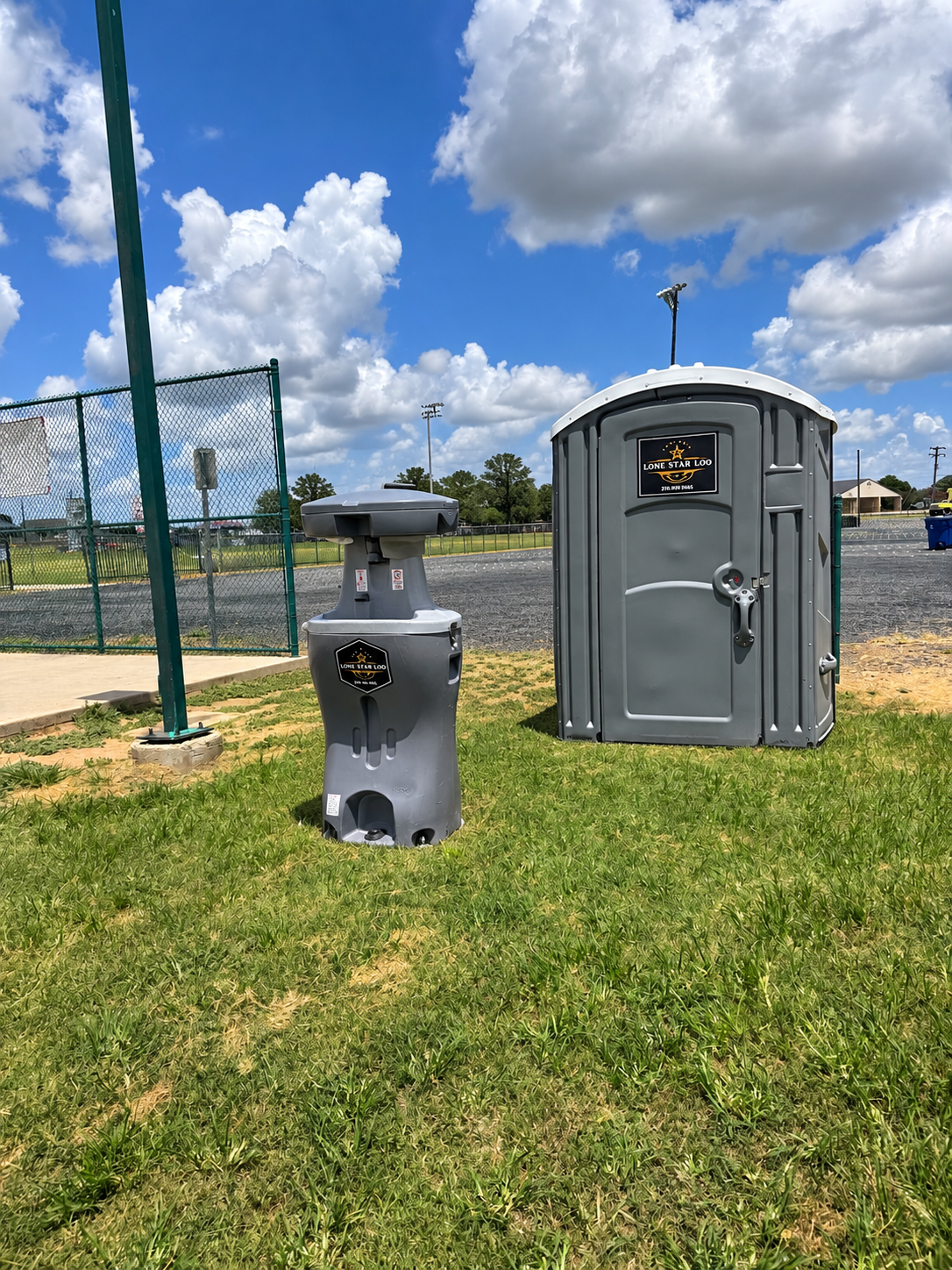 Gray portable toilet and outdoor sink on grass beside a fenced field under a sunny blue sky
