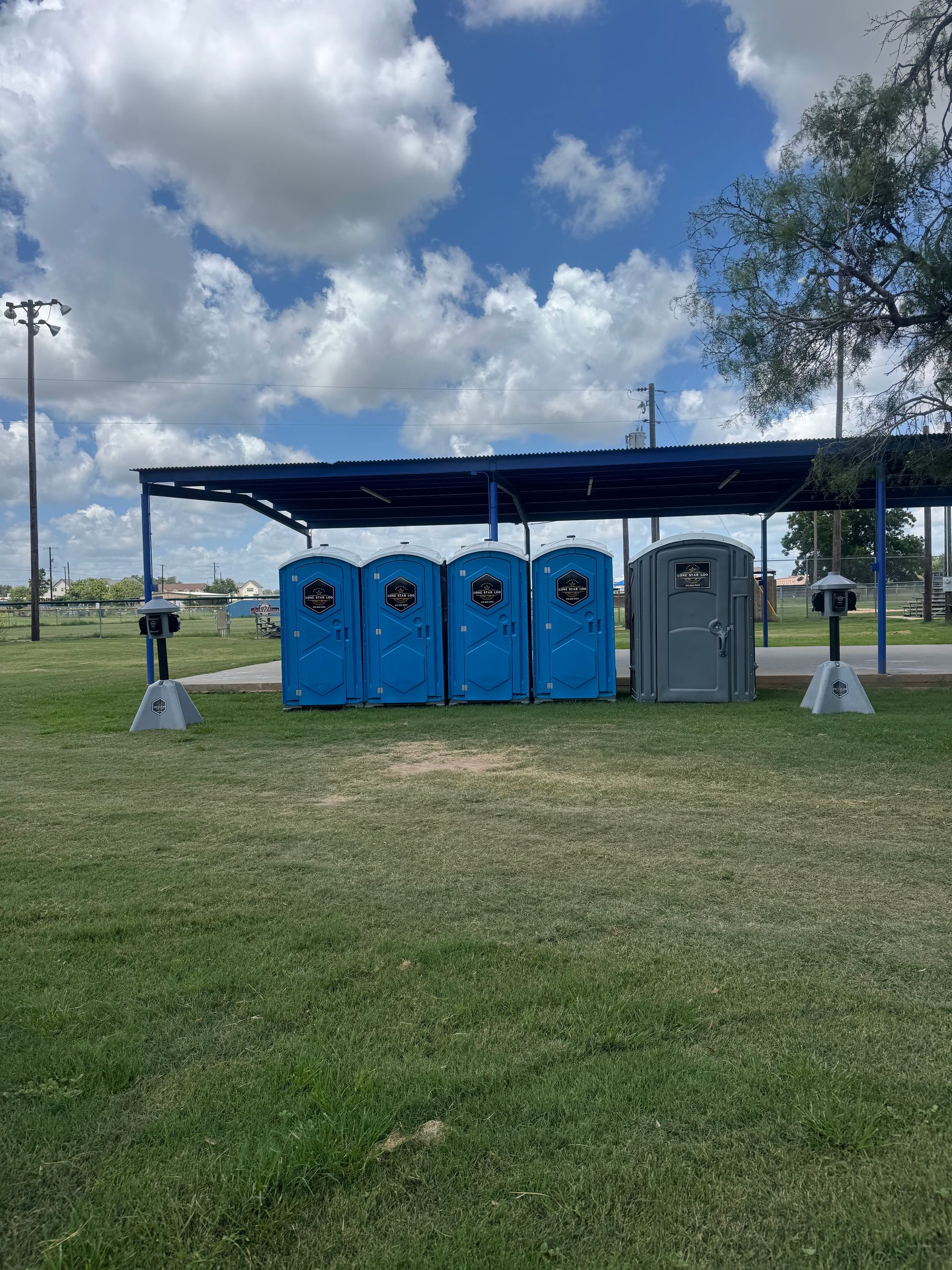 Portable toilets under a blue canopy in a grassy field on a partly cloudy day