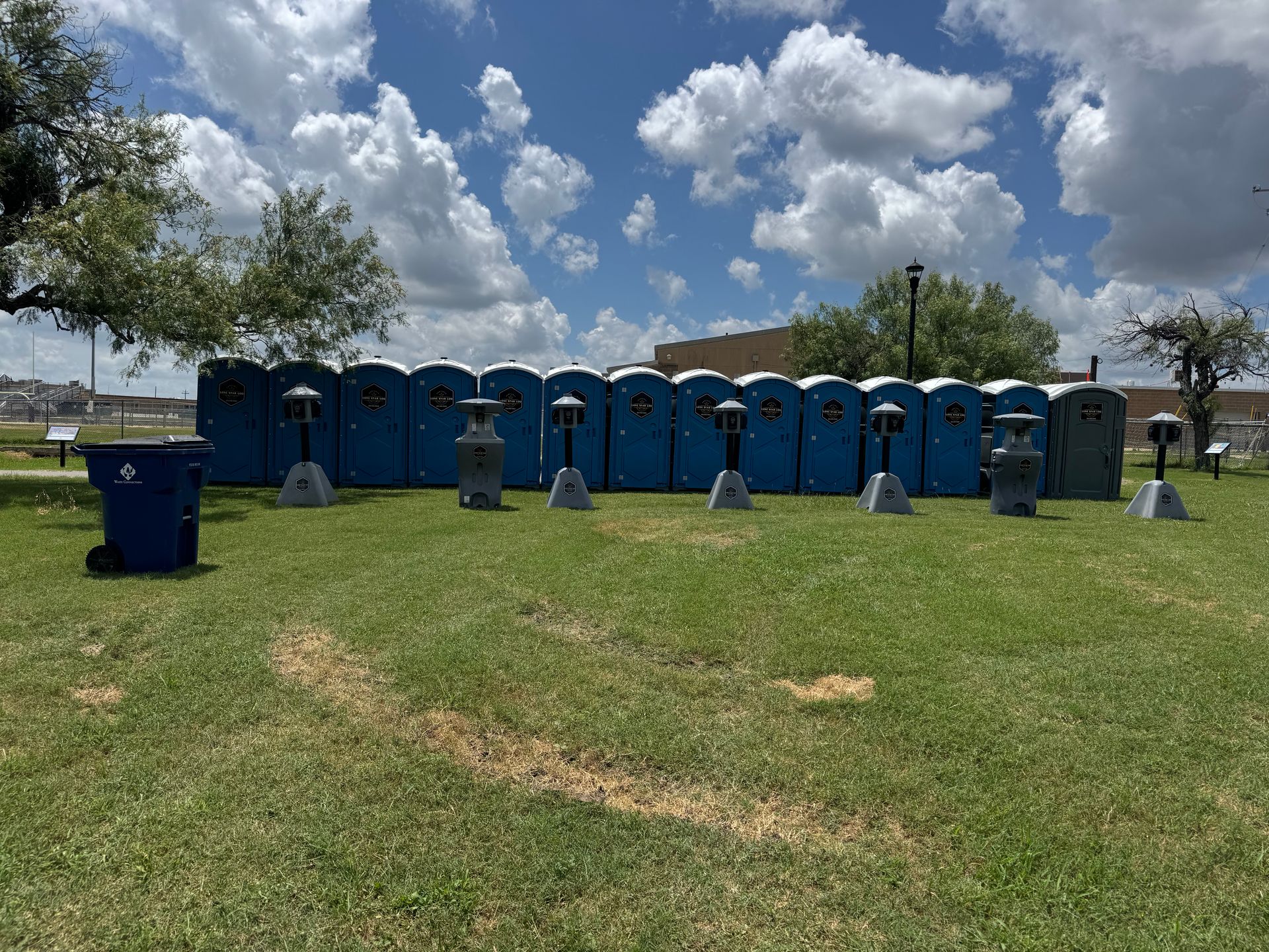 Row of blue portable toilets on a grassy field under a partly cloudy sky, with one trash bin in front