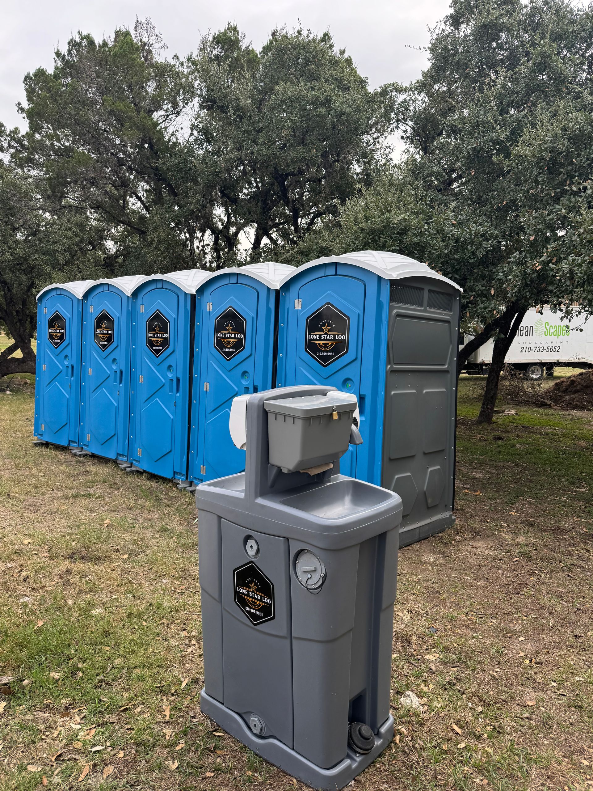 Gray and blue portable toilets lined up outdoors on grass near trees.