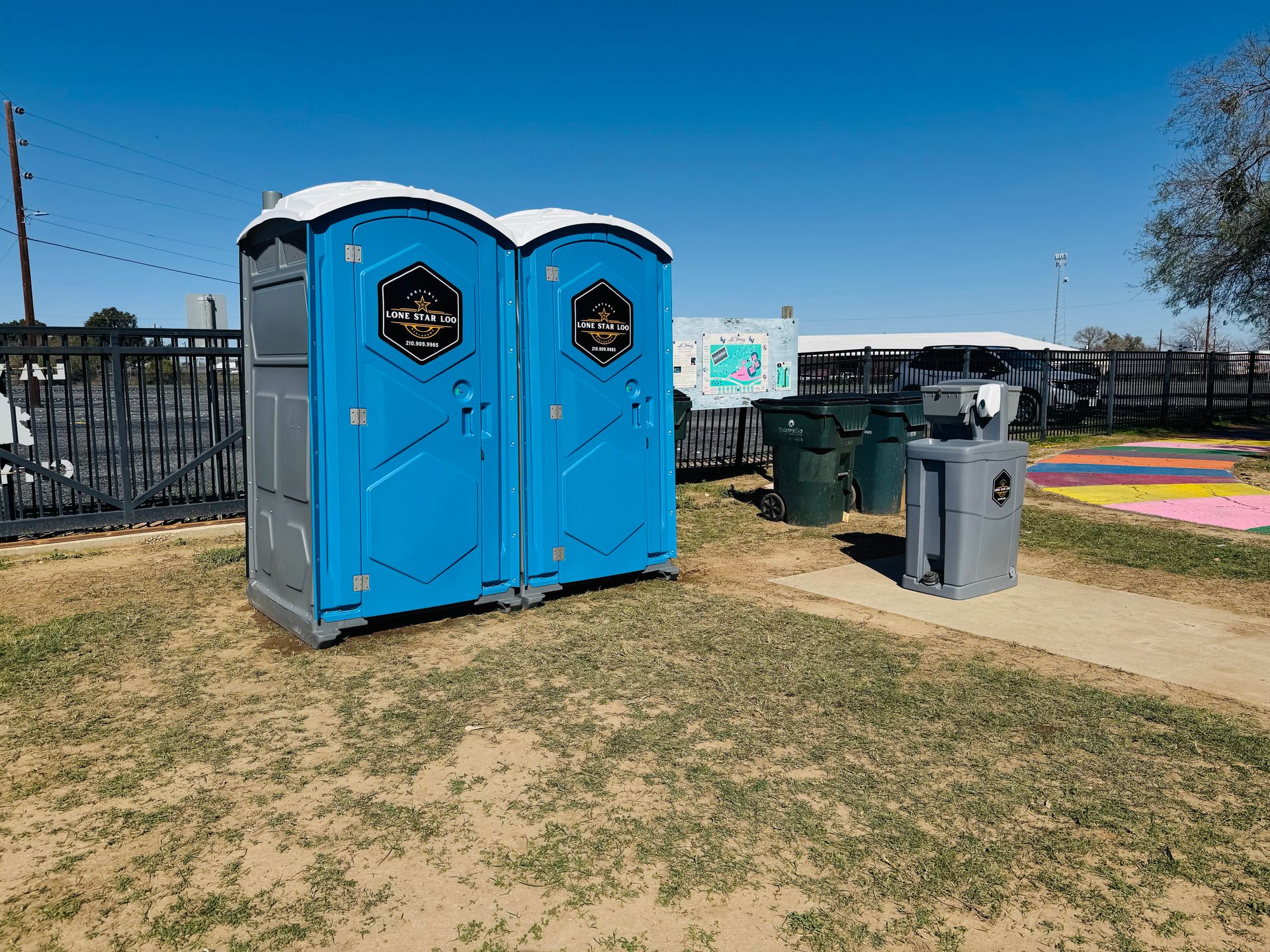 Two blue portable toilets on a grassy path beside a trash can under a clear sky