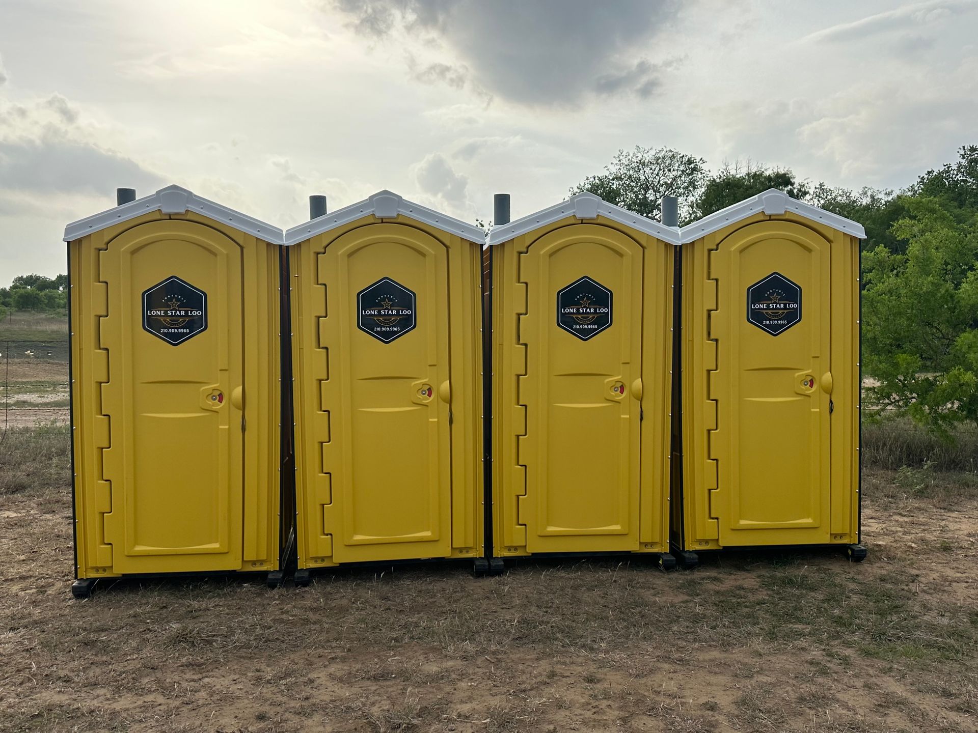 Four yellow portable toilets lined up on a dirt field under a cloudy sky.