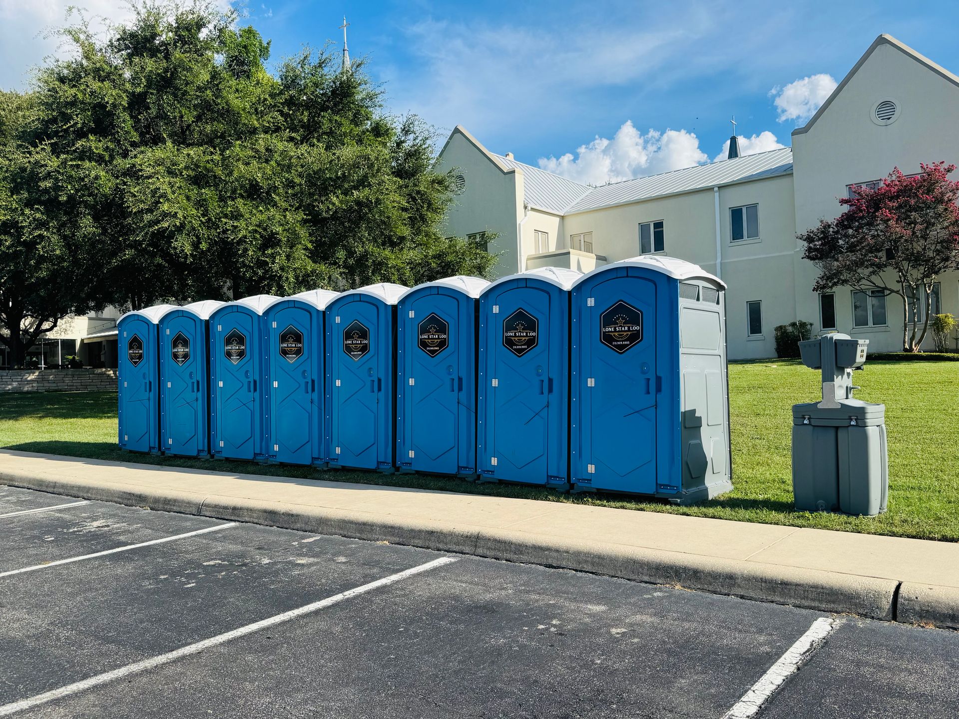 Row of blue portable toilets beside a parking lot and apartment buildings on a sunny day