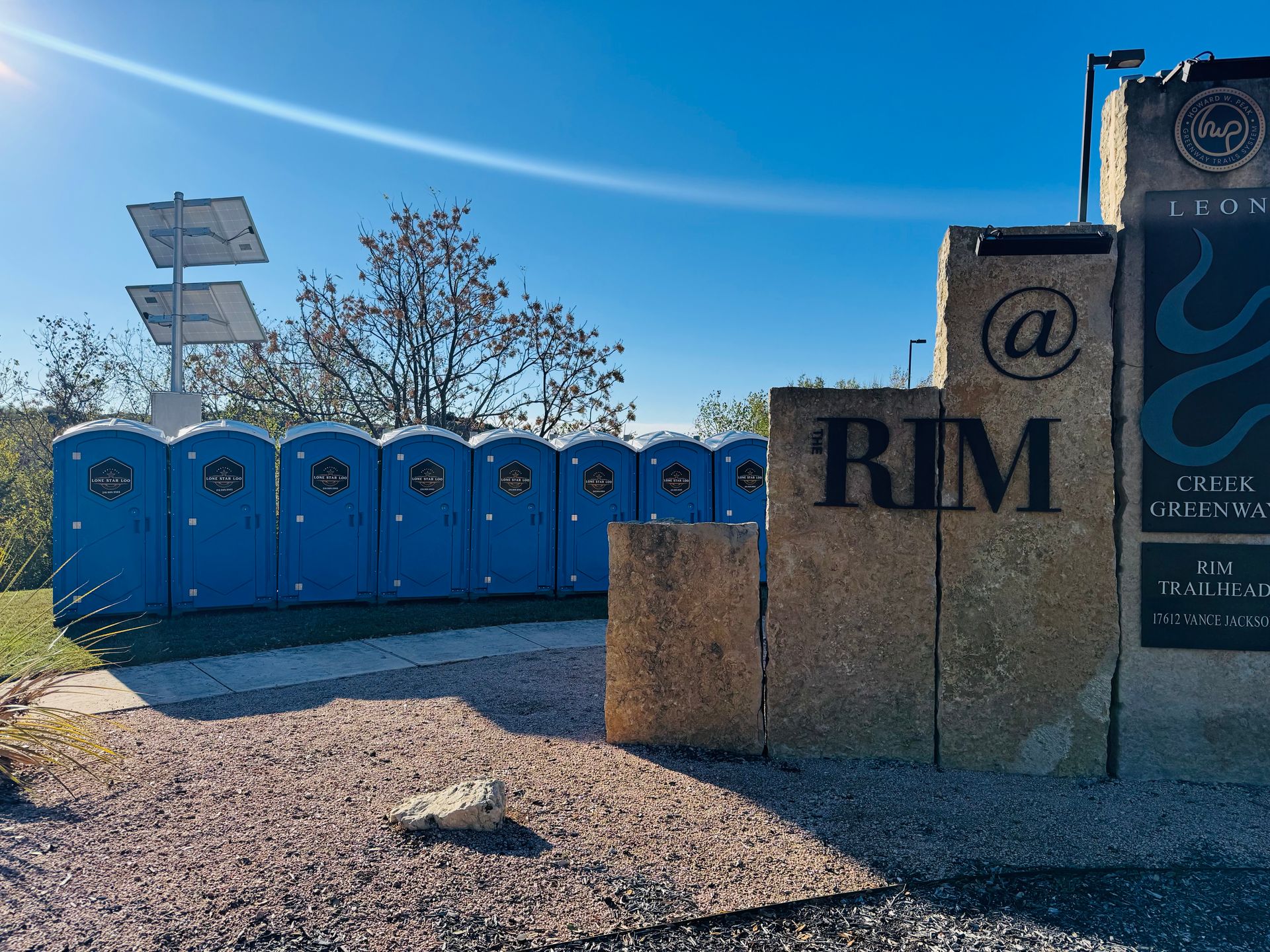 Row of blue portable toilets beside a stone sign in a sunny gravel lot with a desert backdrop