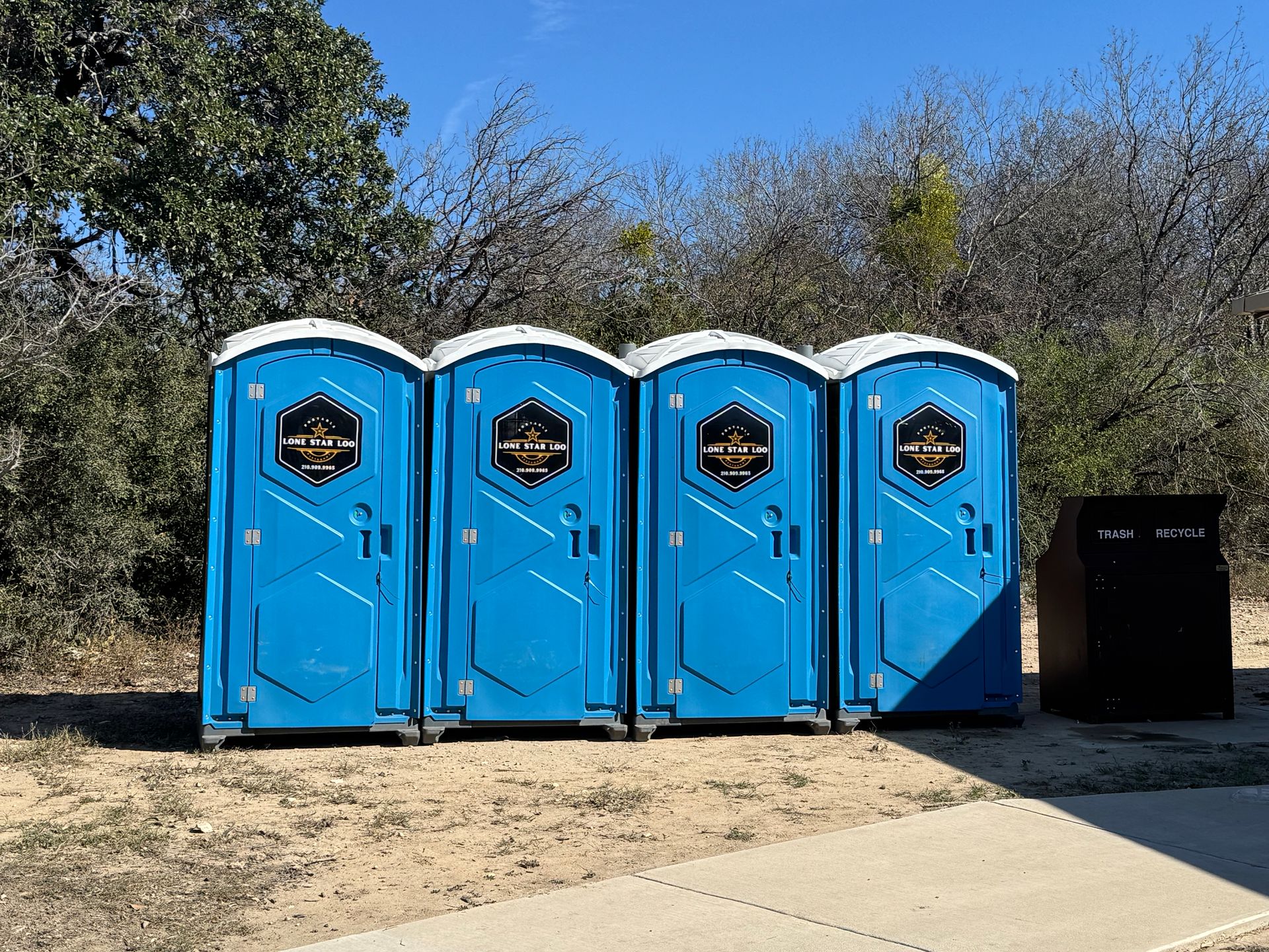 Four bright blue portable toilets lined up outdoors beside a paved path and bushes.