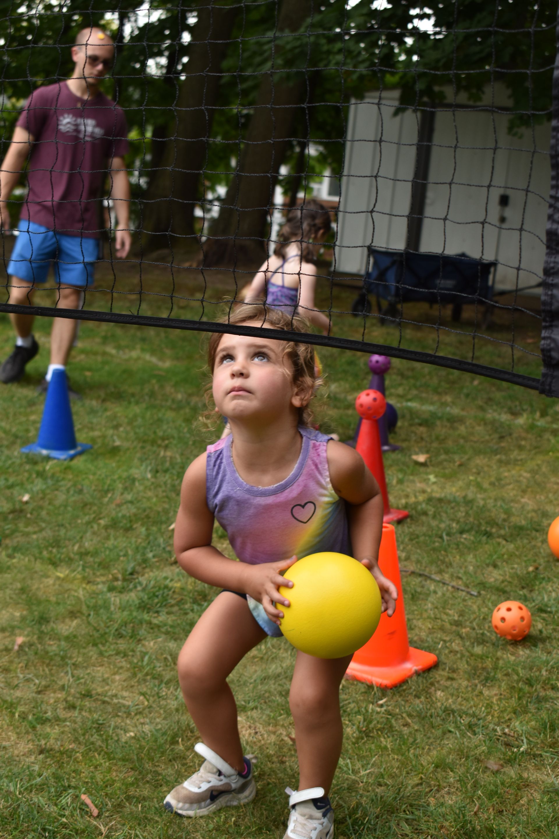 A little girl is holding a yellow ball in front of a volleyball net.