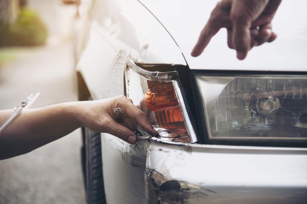 Hands pointing at damage on the front of a white car, possibly after an accident.