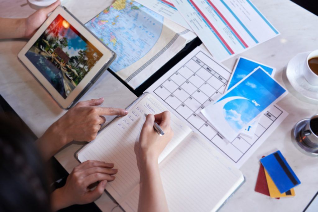 Hands planning travel itinerary on a desk: tablet, notebook, calendar, map, coffee, credit cards.
