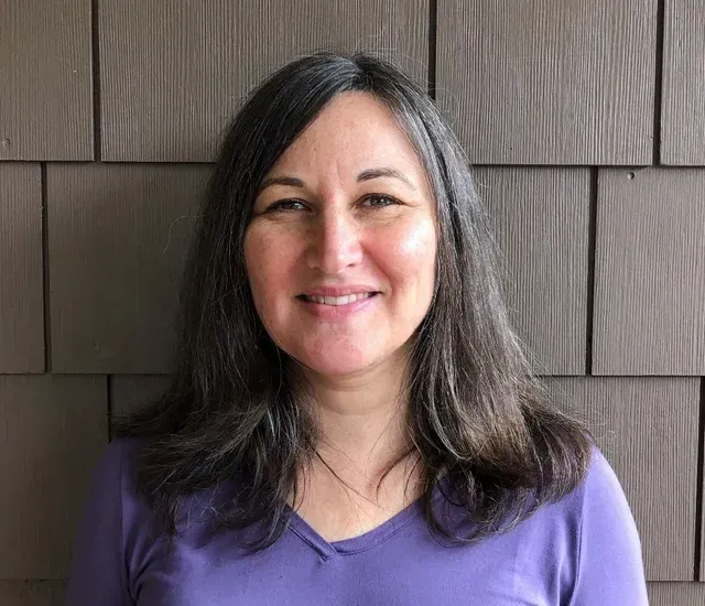 A woman in a purple shirt is smiling in front of a brown wall.