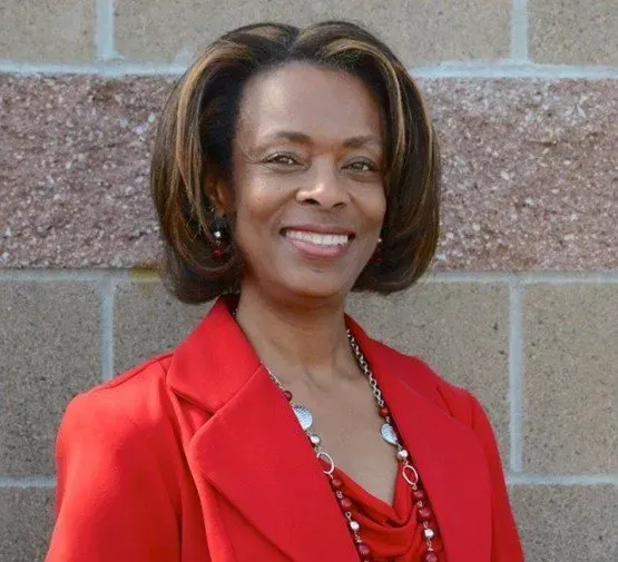 A woman in a red jacket and necklace is smiling in front of a brick wall.